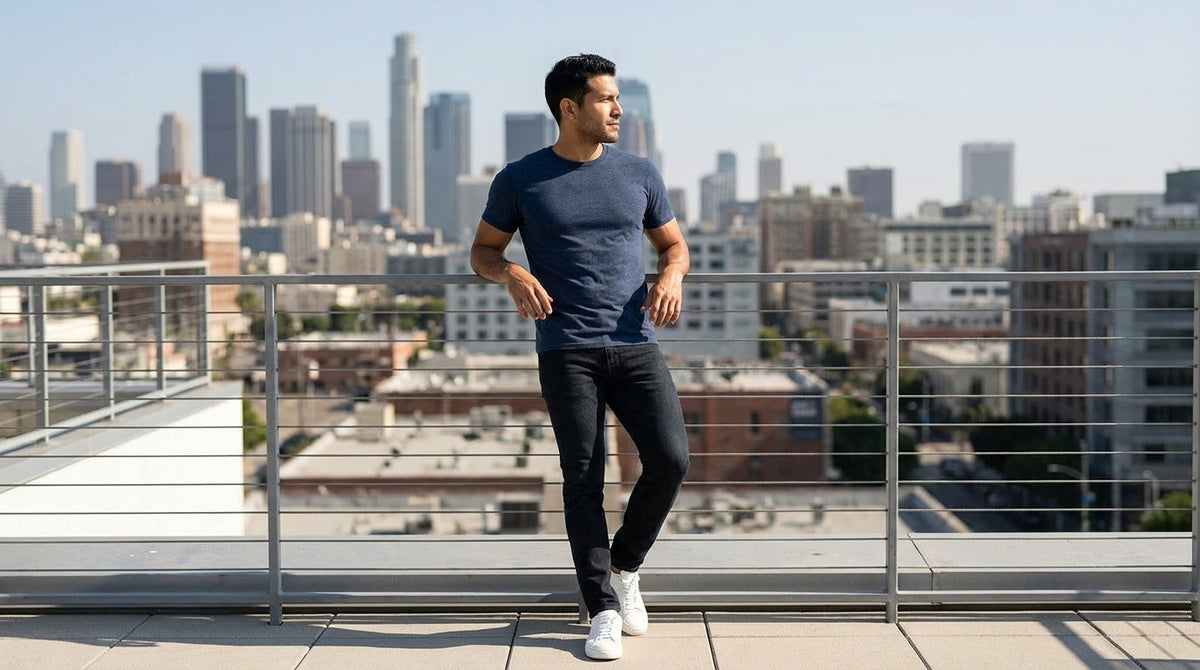 Man in a navy crew neck tee leaning on a rooftop railing above the city skyline