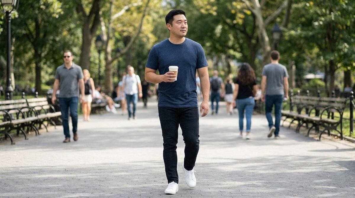 Man in a navy crew neck tee walking through a city park with a coffee
