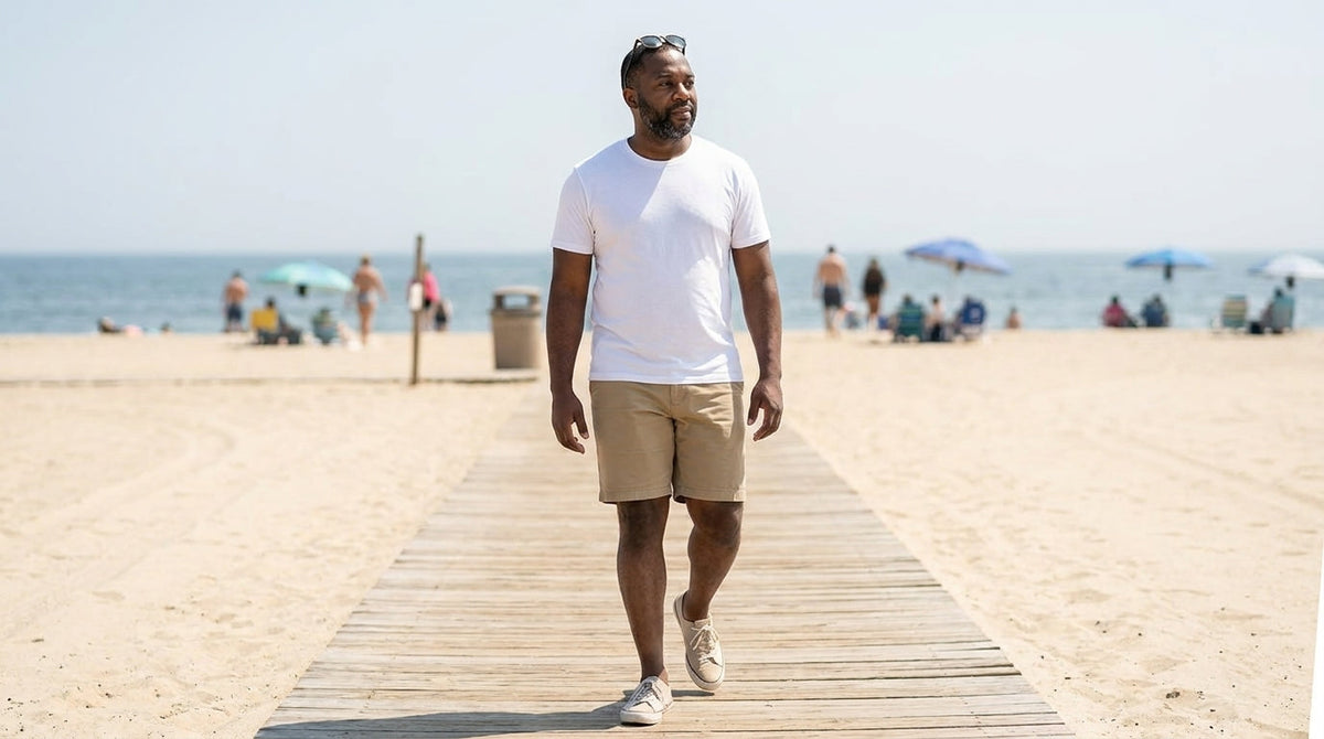Man in a white crew neck tee and khaki shorts walking on a beach boardwalk