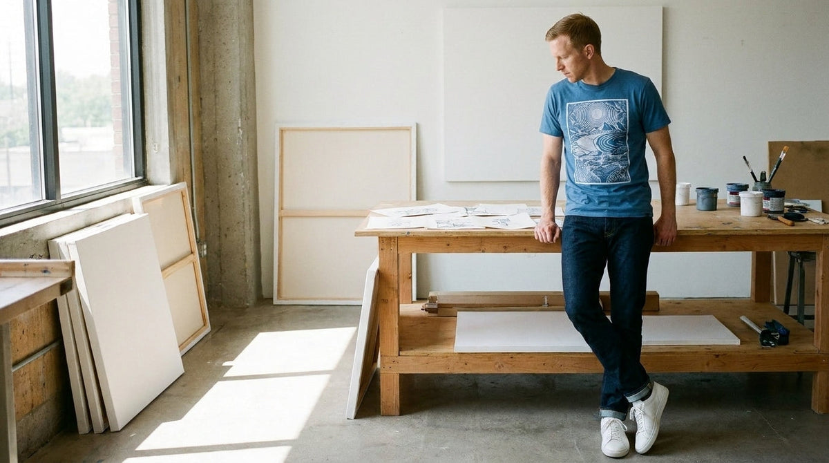 Man in an INTO THE AM graphic tee leaning over sketches in an art studio